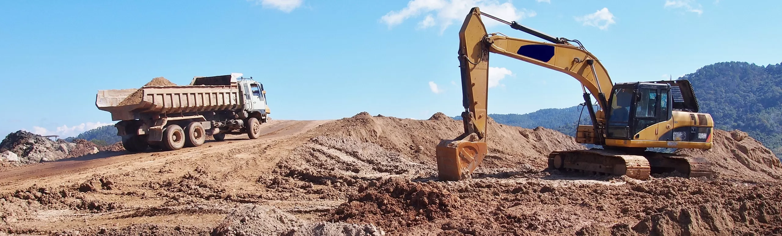 Open mining pit with an excavator sitting idle while a dump truck drives  out of the pit with dirt in the back of the truck